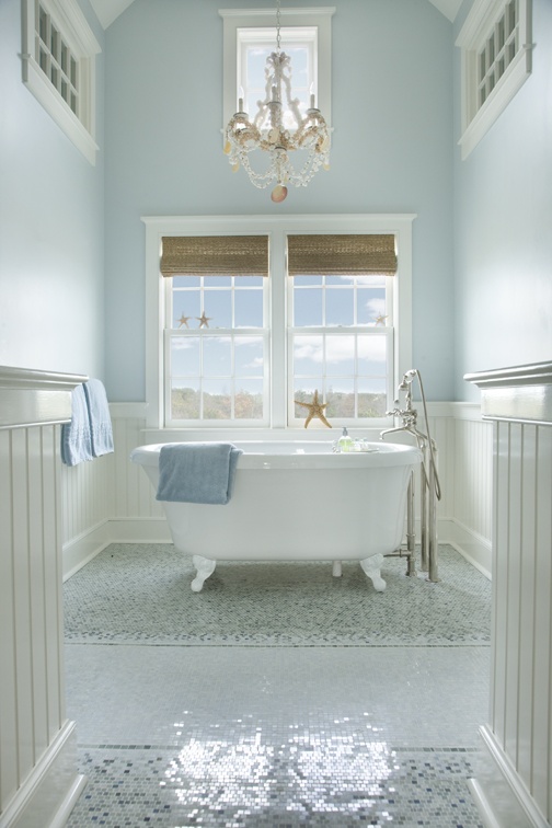 a light blue and creamy bathroom with a mosaic floor, woven Roman shades, a clawfoot bathtub and a chandelier