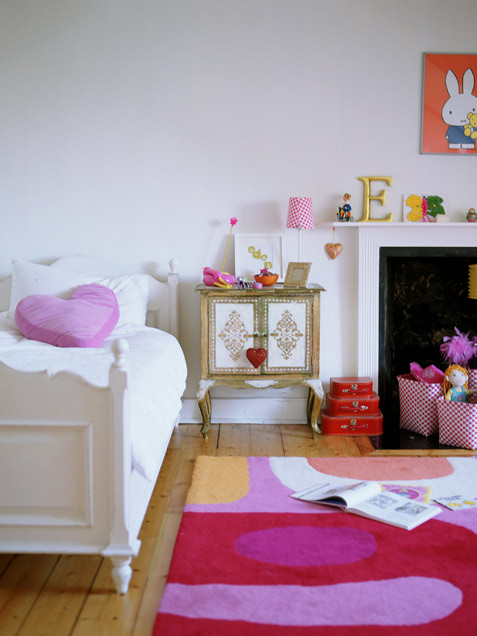 A colorful and whimsical girl's room with a non working fireplace, a bed, a pink heart pillow and a bold rug, a whimsical inlay cabinet and bold and colorful decor