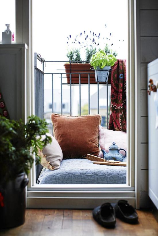 a tiny balcony with a cushion on the floor and some pillows, potted greenery and blooms and a blanket to have a cup of tea