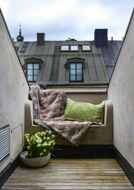 A small attic balcony with a built in upholstered bench, pillows and a blanket and a potted flower is a cool space to get some sunshine