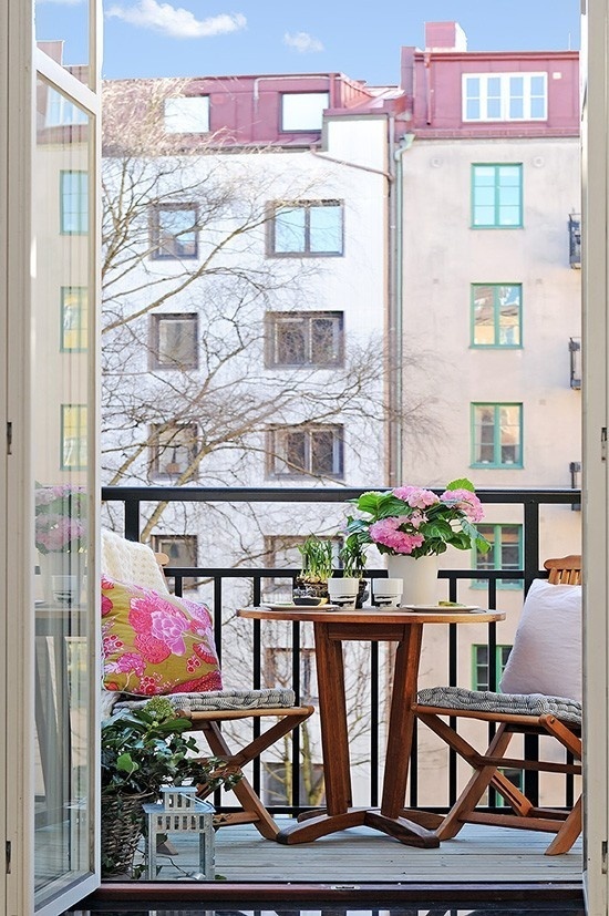 a small and lovely balcony with wooden folding furniture, some pillows and blankets and potted greenery and blooms