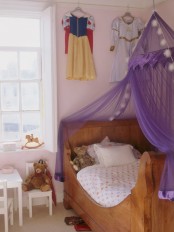 a light pink girl’s room with a stained bed and a purple canopy over it, string lights, play furniture and some princess dresses