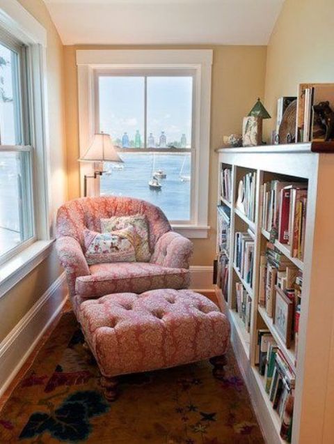 a sunroom turned into a reading nook with a large bookshelf and a pink lounge chair plus a lamp