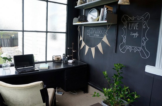 a small moody home office with a chalkboard accent wall, a black desk and a neutral chair, wall-mounted shelves and potted plants