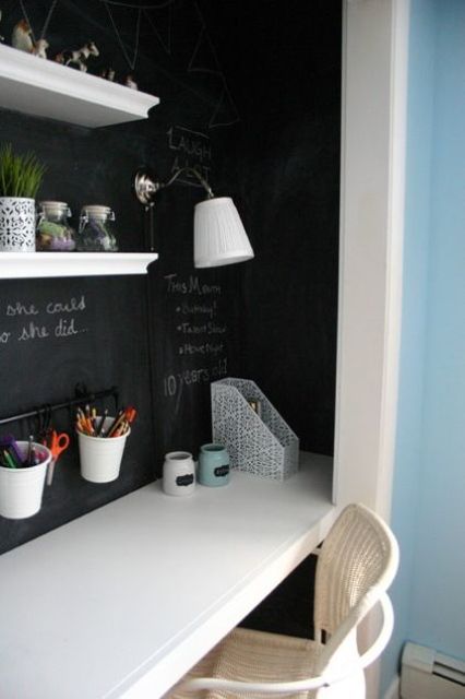 A small built in working nook with a chalkboard, wall mounted shelves, a white desk and a chair, a wall lamp
