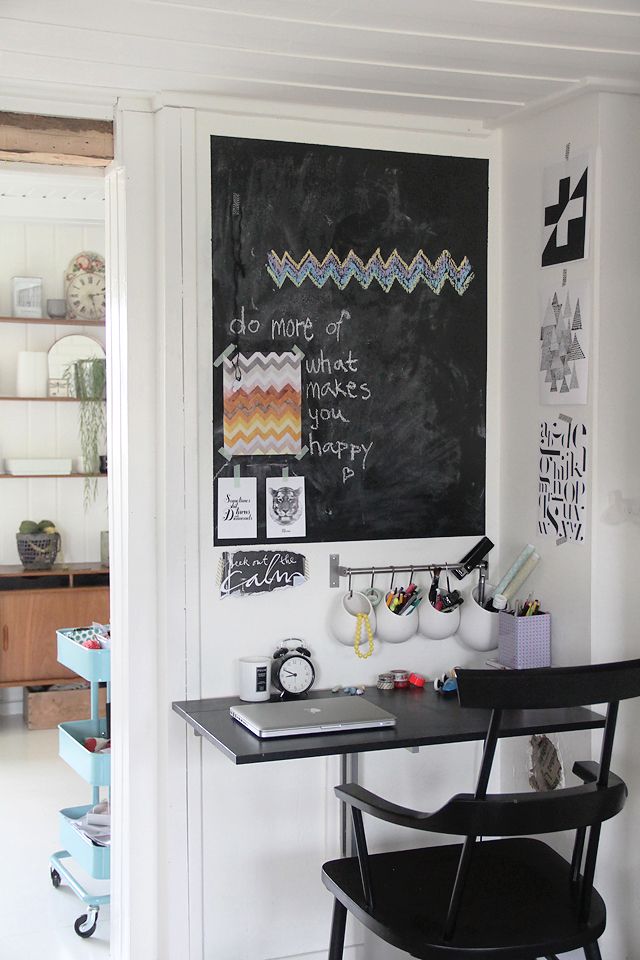 A tiny working studying space with a small wall mounted desk, a black chair, a chalkboard and railing with various organizers is a lovely space