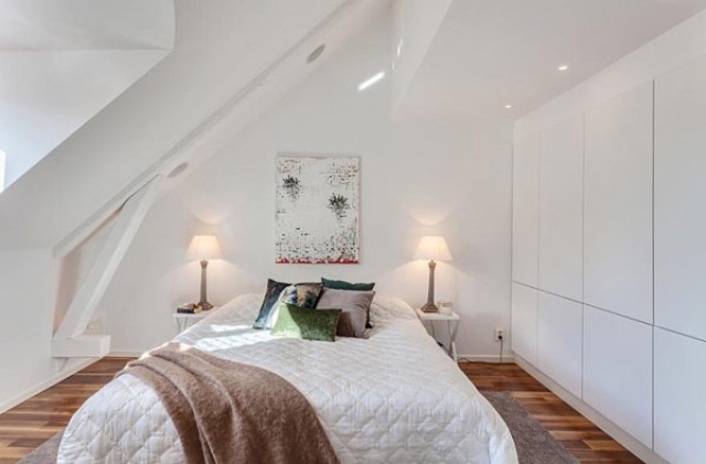 A tiny white bedroom with sleek white cabinets, built in lights, a bed and lamps and much natural light