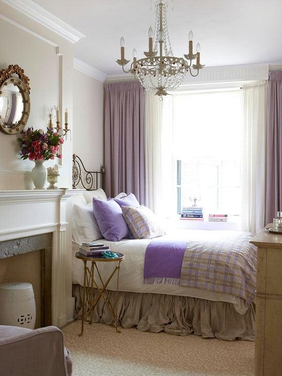 A small vintage bedroom with a crystal chandelier, purple curtains and bedding, a non working fireplace and a mirror