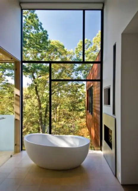 A modern bathroom with a glazed wall, a built in fireplace, an oval tub and a gorgeous view of the fall forest