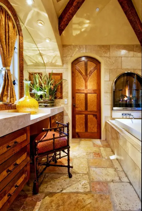 A vintage bathroom clad with stone, with a wooden vanity, a metal chair with red upholstery, a tub clad with tiles and a built in fireplace in the wall over it
