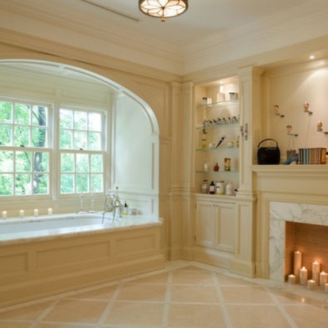 A creamy vintage bathroom with a tub placed by the window, with built in shelves, a fireplace and lots of candles for a chic and stylish look