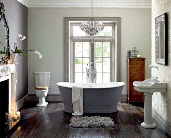 A vintage bathroom with a dark floor, a vintage fireplace with candles, a grey bathtub, a free standing sink and a crystal chandelier