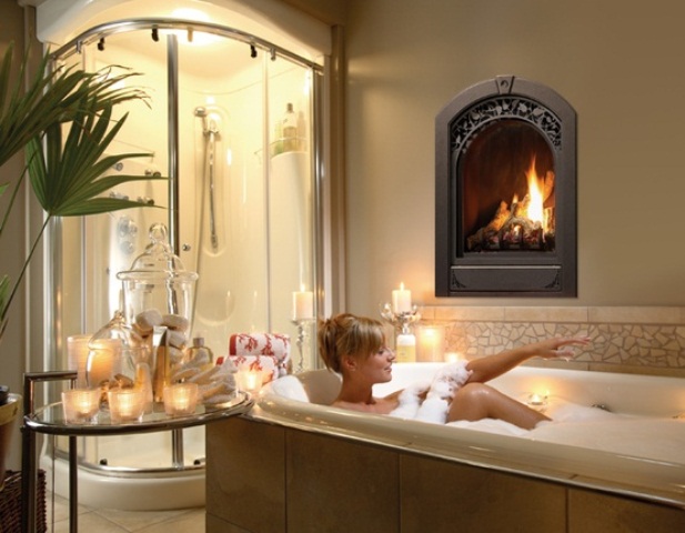 A refined bathroom with a lit up shower, a built in fireplace, a bathtub clad with large scale tiles and a glass table with candles