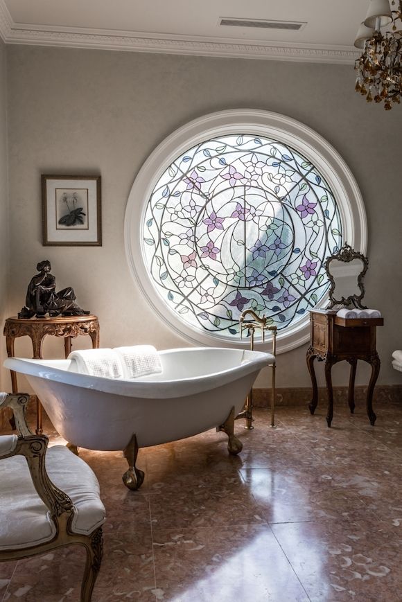 A fantastic and jaw dropping vintage inspired bathroom with a round window with stained glass, a clawfoot bathtub and vintage carved furniture just wows
