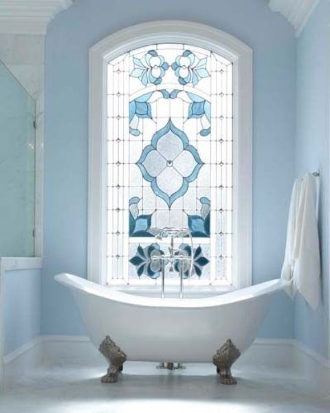 An airy and light filled serenity blue bathroom with a shower space, with a large stained glass window and a vintage clawfoot bathtub