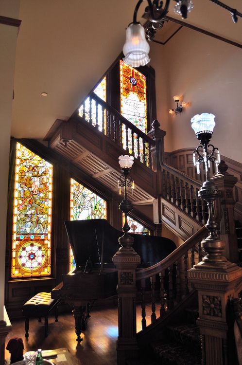 A refined vintage space with a dark stained carved staircase, with a piano and beautiful lamps, with stained glass windows that keep privacy and add color