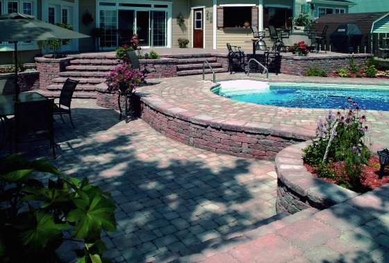 A kidney shaped pool with a stone deck plus an additional raised touch, greenery and blooms and an outdoor dining set