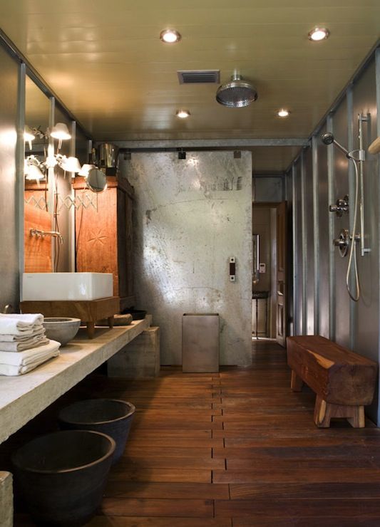 a modern industrial bathroom with steel and concrete walls, a wooden floor, a long concrete vanity, stone bowls, a bench is a statement space