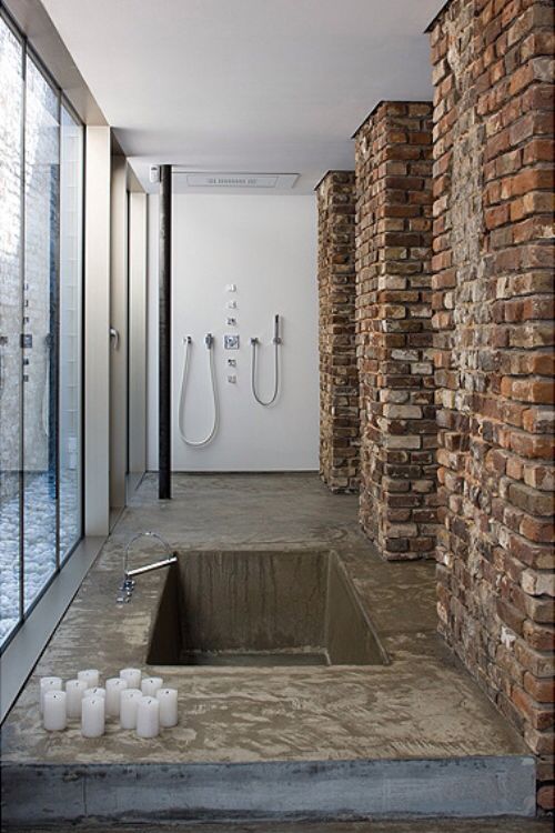 a modern industrial bathroom with a concrete floor, brick pillars, a glazed wall, a built-in tub and candles around it
