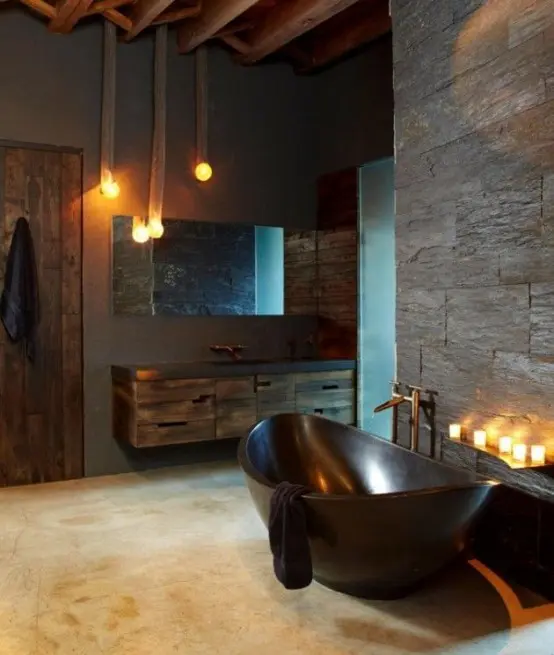 a mascauline meets industrial bathroom with concrete and faux stone walls, rough wooden furniture and a door, a polished stone bathtub and candles and pendant lamps