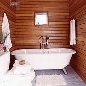 a small bathroom clad with rich stained wood, with an elegant vintage bathtub and white textiles and a toilet