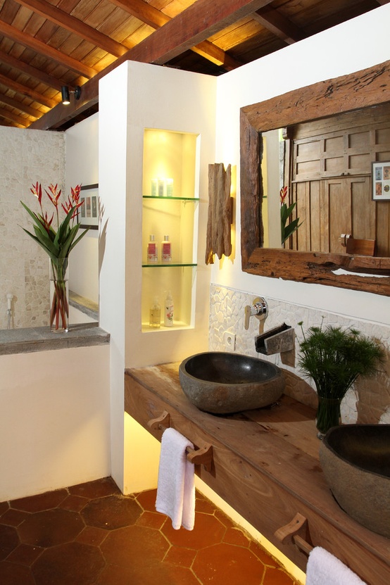 A tropical bathroom with a built in wooden vanity, stone sinks, built in niches for storage, a mirror in a rough wooden frame