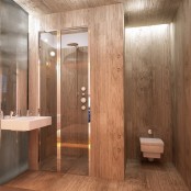 a contemporary bathroom fully clad with stained wood, with a shower enclosed in glass and wood, with a wall-mounted sink and a modern toilet