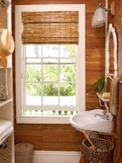 a small rustic bathroom fully clad with wood, with a window with shades, a sink, some baskets for storage and a large storage unit