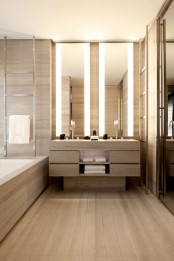 a minimalist bathroom fully clad with light stained wood, with a large double vanity and lit up mirrors, a large bathtub clad with wood