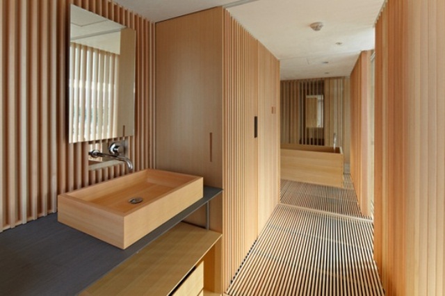 A minimalist light stained wooden bathroom with a wood slab wall and floor, with large storage units and a wooden bathtub