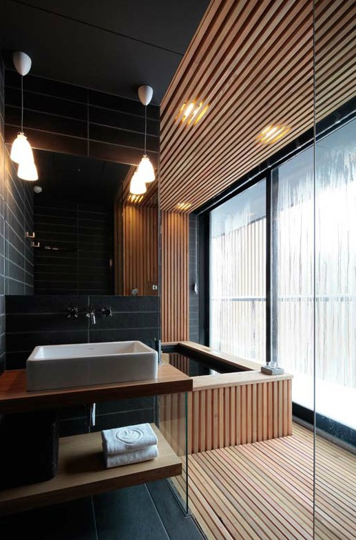 a minimalist bathroom done with light stained wooden slab and black skinny tiles, with pendant lamps and a large window for more light