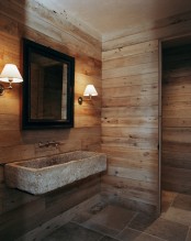a rustic bathroom clad with light stained wood and with tiles on the floor, with a wall-mounted stone sink and sconces