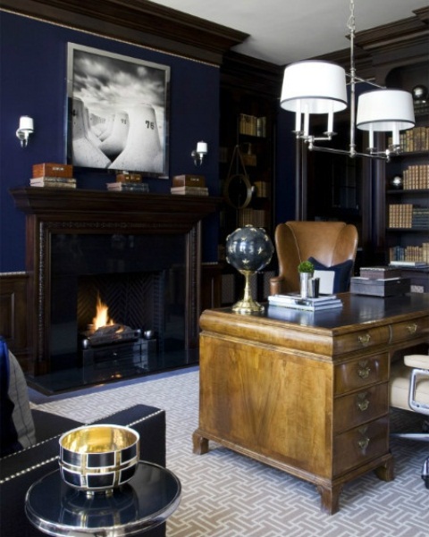 An elegant vintage inspired masculine home office with navy walls, dark bookcases, a light stained desk and a leather chair