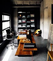 a moody home office with concrete walls and a floor, a black bookcase, a wooden desk with various levels plsu a leather chair
