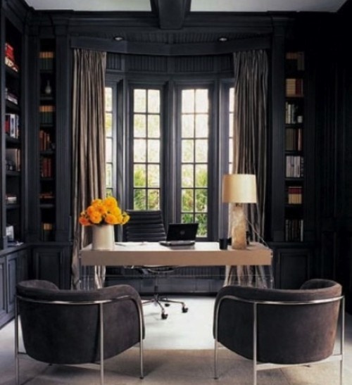 A moody home office with graphite grey walls and built in bookcases, a white desk and comfy upholstered chairs