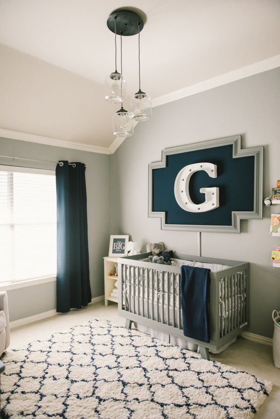a lovely nursery with a grey crib with neutral bedding, a printed rug, black curtains, a monogram artwork, a cluster of pendant lamps