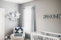 a monochromatic nursery with grey walls and white furniture, black and white textiles and a rug, a wall mounted shelf and a name over the bed