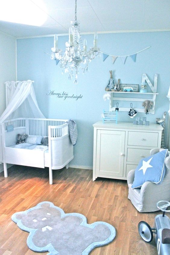 A dreamy light blue nursery with vintage white furniture, a grey chair with pillows, a rug and a wall mounted shelf plus a lovely crystal chandelier