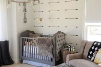 an eclectic nursery with an arrow accent wall, a grey crib with a tufted detail, a taupe chair and printed textiles, a pouf is a chic space