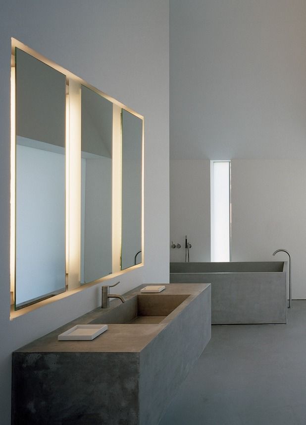 An ultra minimalist concrete bathroom with a concrete bathtub and a floating vanity with a built in sink plus mirrors with built in lights