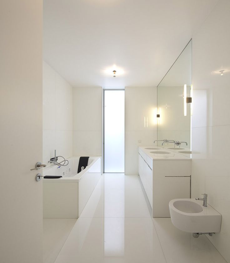A white minimalist bathroom with large scale white tiles all over, a long double vanity and white appliances plus a vertical window with frosted glass
