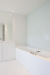 a white sleek minimalist bathroom with a glass wall, a statement mirror, white furniture and appliances