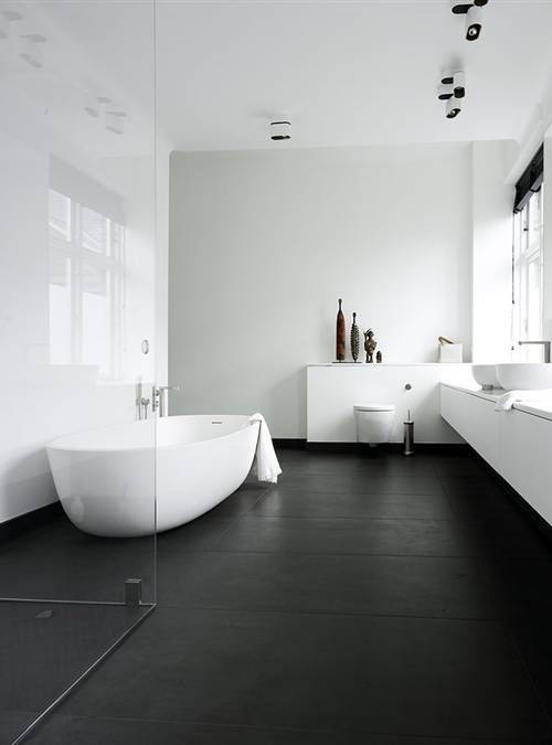 a contrasting bathroom with a black floor,  white walls, a long white vanity, several windows and black Roman shades, a seamless glass enclosed shower