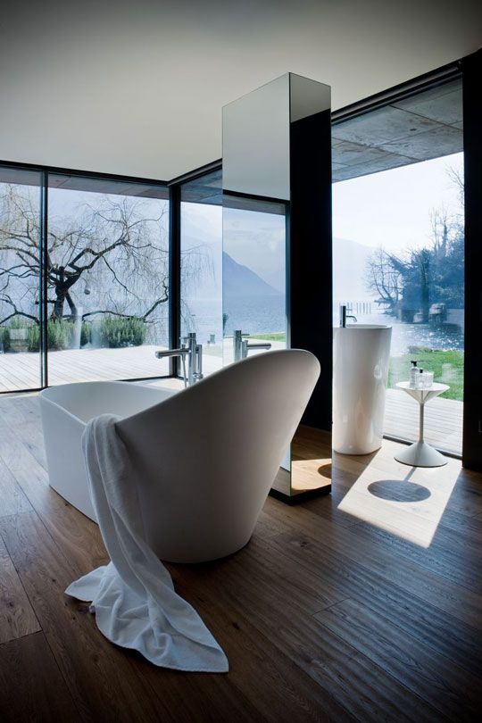 A chic glass enclosed minimalist bathroom with a wooden floor, white free standing appliances and a mirror pillar