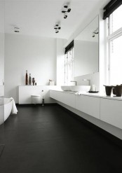 a contrasting minimalist bathroom with a black floor, a long white vanity, a statement mirror, two windows and white furniture and appliances