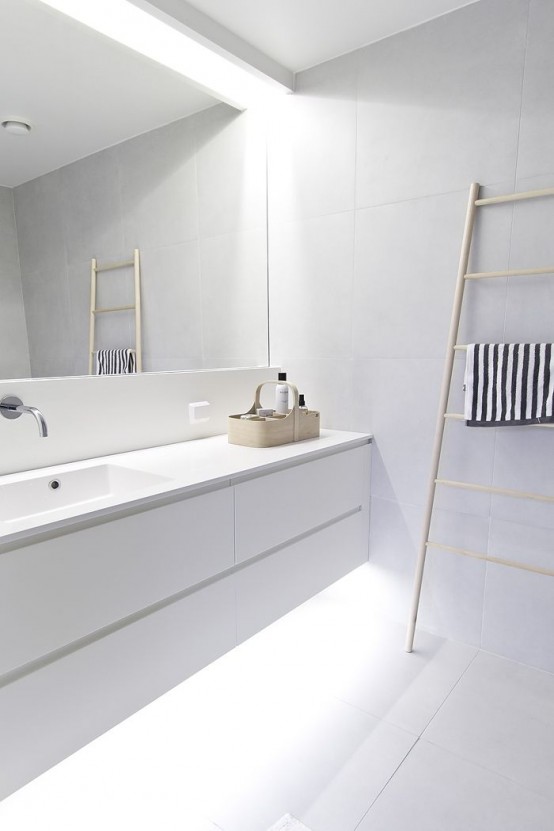 an ultra-minimalist white bathroom with a floating vanity with built-in lights, a statement mirror and a ladder for storing towels