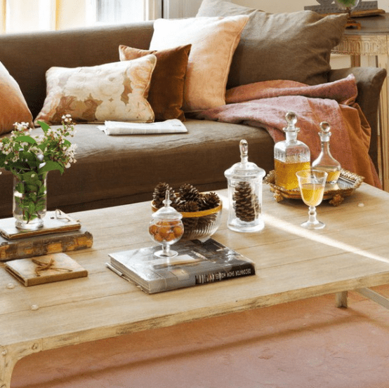 a light-colored wooden coffee table with pinecone arrangements, books, greenery and alcohol in chic glassware