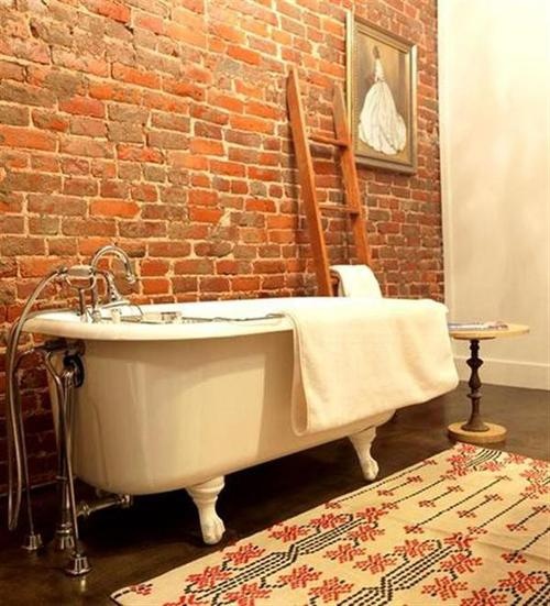 A vintage inspired bathroom with a red brick wall, a colorful rug, a ladder, a side table and an artwork