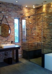 a contemporary bathroom with red brick walls, a glass shower space, a dark vanity and a large round mirror