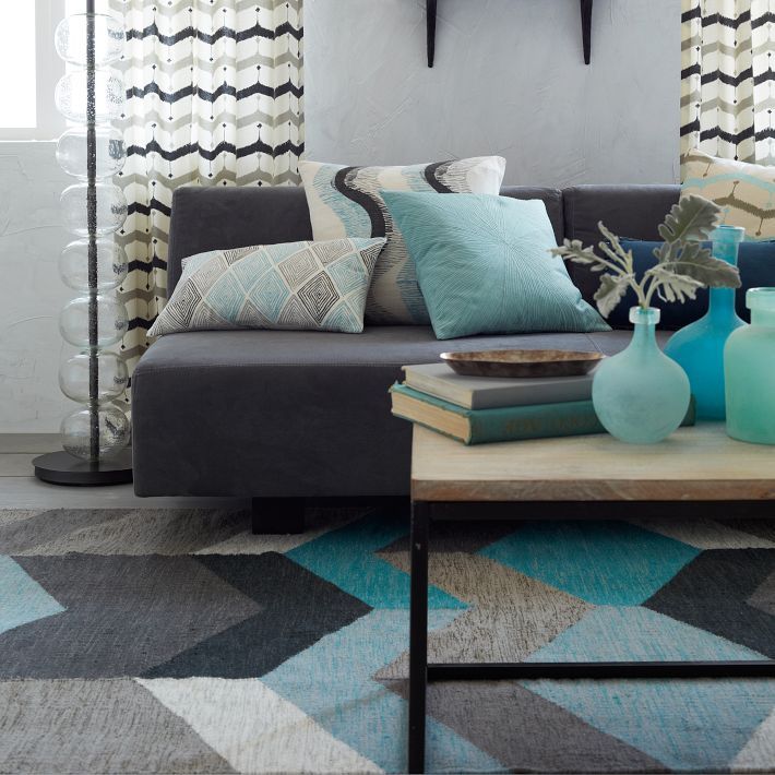 A mid century modern living room with a chevron printed rug, a coffee table with blue glass bottles, a grey sofa with printed pillows and chevron print curtains